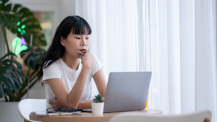 Concentrated Asian woman working by checking data on laptop Serious thinking and planning, smartphone, coffee and documents on the table.