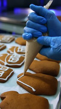 Confectionery worker holding a pastry bag and squeezing white cream on the cookies. Gingerbread houses close up. Trays with pastry in blur at background. Vertical video