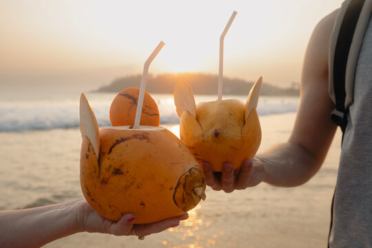 Man and woman drinking fresh coconut water together on idyllic sand beach. Healthy natural refreshment at sunset. Mirissa in Sri Lanka..