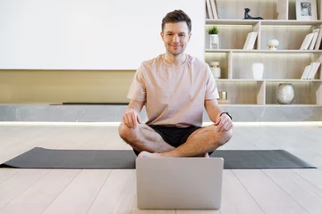 Foto auf Acrylglas A serene man meditates in lotus position on a yoga mat with a laptop before him, integrating mindfulness and technology in a modern home. © muse studio