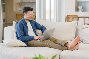 A relaxed man with a pleasant smile works on a laptop, sitting barefoot on a sofa in a light, contemporary living space.