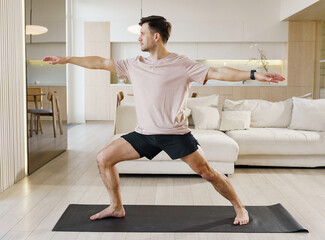 A man practices the Warrior II yoga pose with precision and calmness, harmonizing his body and mind in a well-lit, open home space.
