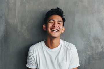 Joyful young man laughing with eyes closed, leaning against a gray background, wearing a white t-shirt, embodying happiness and positivity.