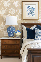  A bedroom with blue and white floral wallpaper, beige walls, a vintage wooden bedside table with a marble top, and an elegant lamp on the nightstand. The room is adorned with navy blue pillows.