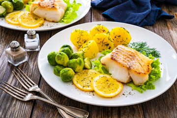 Fried cod loin, boiled potatoes and cooked Brussels sprouts on wooden table
