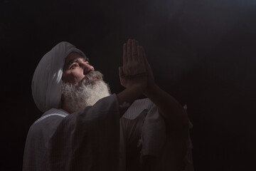 A senior man in a turban is associated with a Muslim standing in dark smoky background light ray...