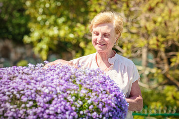 Elderly woman standing near a large flower pot with purple flowers and caring for plants