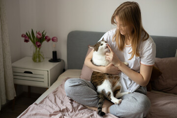 Young blonde 30s woman is resting with a Stripped White cat on the bed at home one spring day. Morning at home. Cute cat licking fingers of female owner in daytime at home. Feeding.