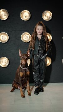 Little girl and Doberman at photo shoot in studio. Girl in stylish suit with Doberman on background of wall with round lamps. Concept of style, friendship, photo art