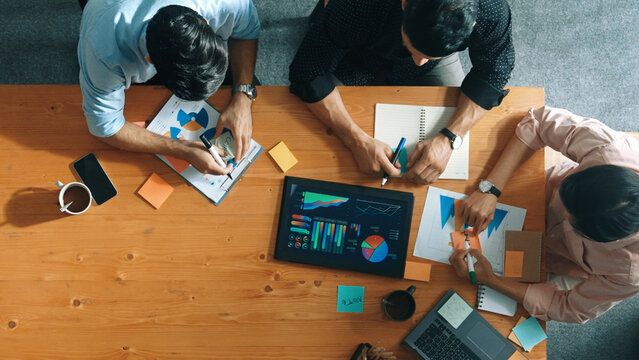 Executive Manager Explain Financial Graph To Colleague At Meeting Room. Top Aerial View Of Diverse Marketing Team Sharing Idea And Planning Strategy While Sitting At Table With Tablet. Convocation.