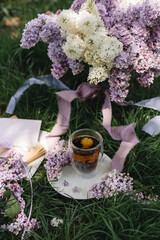 Serving on a green lawn: a lilac in a vase, a floral Chinese natural tea brewed in a glass cup, a card and purple ribbons. Tea party on a picnic. Romance