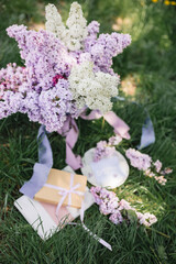 A greeting card with the inscription "Bonjour" lies on the grass, near a bouquet of purple, white and soft blue lilac and a purple silk ribbon. Decor © Olga
