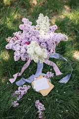 A greeting card with the inscription "Bonjour" lies on the grass, near a bouquet of purple, white and soft blue lilac and a purple silk ribbon. Decor © Olga