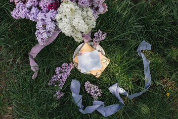 A greeting card with the inscription "Bonjour" lies on the grass, lilac flowers and a purple silk ribbon. Top view © Olga