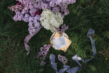 A greeting card with the inscription "Bonjour" lies on the grass, lilac flowers and a purple silk ribbon. Top view © Olga