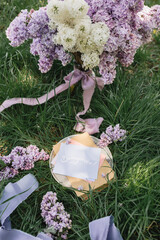 A greeting card with the inscription "Bonjour" lies on the grass, lilac flowers and a purple silk ribbon. Top view © Olga