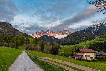 Village road in Alpine meadow and buildings against the Geisler mountains covered with snow at sunset, Val Di Funes valley in Dolomites mountains, South Tyrol, Italy. Italian Alps in springtime - obrazy, fototapety, plakaty