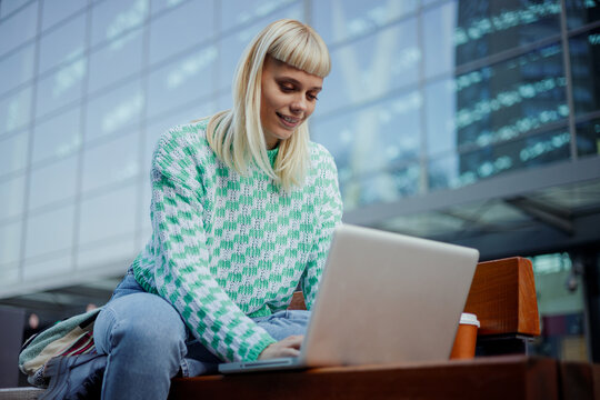 An e-nomad is sitting on street downtown and typing on a laptop.