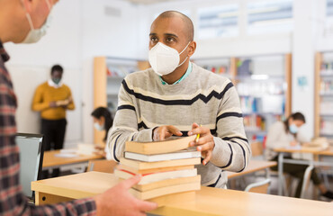 Adult hispanic student in protective face mask talking to librarian while returning books to...