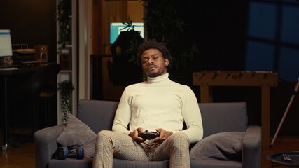 African american man in living room playing video games on gaming console from couch during leisure...