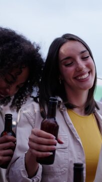 Vertical. Cheerful multiracial young people toast with beer bottles celebrating dancing and laughing on summer outdoors. Group happy millennial friends together enjoying sunset drinks at rooftop party