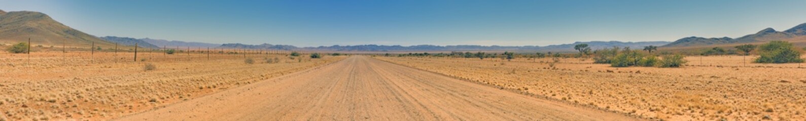 Panoramic picture over a gravel road through the desert like steppe with red sand in southern Namibia under a blue sky