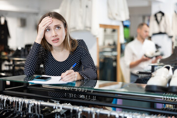 Upset girl store owner makes calculations, checks documents and counts budget deficit using her phone. Female store manager is disappointed about unsatisfactory business profitability results - obrazy, fototapety, plakaty