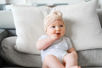 Caucasian female infant dressed in bodysuit and turban on couch, holds hand near mouth. Infant emotions and expressions. - obrazy, fototapety, plakaty