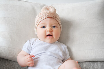 Caucasian female infant dressed in bodysuit and turban on sofa. Disgusted face. Childish emotions...