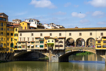 The Ponte Vecchio bridge stands proudly over the Arno River in Florence, flanked by vibrant buildings and reflecting on the water. - obrazy, fototapety, plakaty