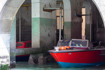 Fototapeta na wymiar A vivid red boat moored in the shadows of an underpass, with the contrasting weathered concrete pillars offering a glimpse into Venice's utilitarian spaces