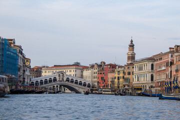 The Rialto Bridge stands proudly over the Grand Canal in Venice, framed by historic facades, under the watchful eye of a campanile in the distance. - obrazy, fototapety, plakaty