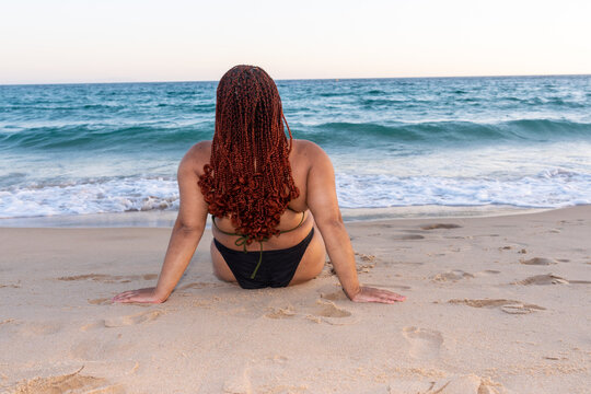 Plus size black girl sitting on the sand on the beach looking at the sea. Young woman with braids on the beach in bikini enjoying her beach vacation.