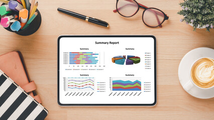 Overhead view of a tablet on a desk showing colorful summary reports, accompanied by office...