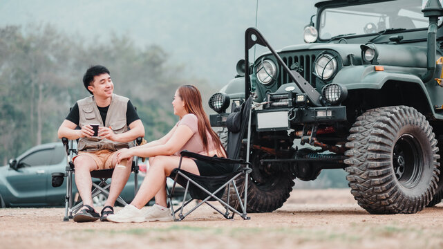 Couple is deep in conversation, enjoying a warm drink while seated beside a rugged off-road car at their scenic campsite.