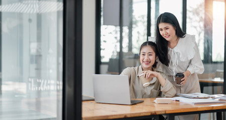 Two smiling female colleagues collaboratively working on a project with a laptop and coffee in a bright office space.