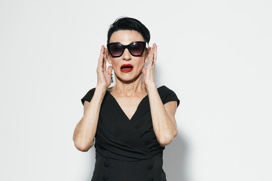 Elegant mature woman in black dress and sunglasses covering her ears while looking at the camera