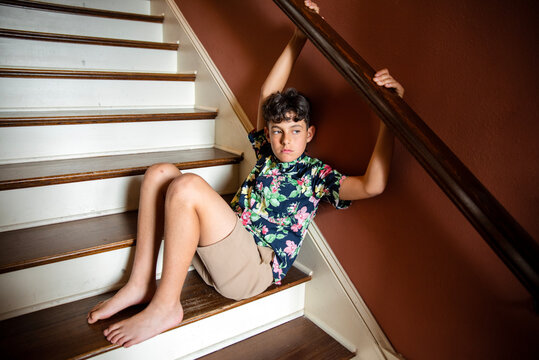 Barefoot preteen boy wearing a floral print Hawaiian shirt and khaki shorts, sitting on a staircase with hands holding the handrail  - Powered by Adobe