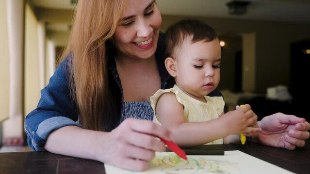 Happy Latin mother and baby daughter having fun painting with crayons at home. Family love relationship concept