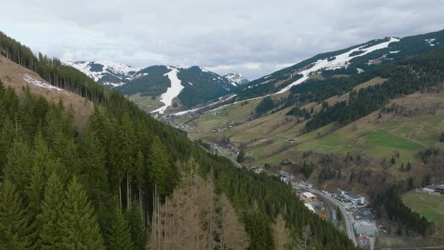 Lush green pines framing a scenic Austrian ski resort in Saalbach-Hinterglemm, traces of snow on slopes, aerial view
