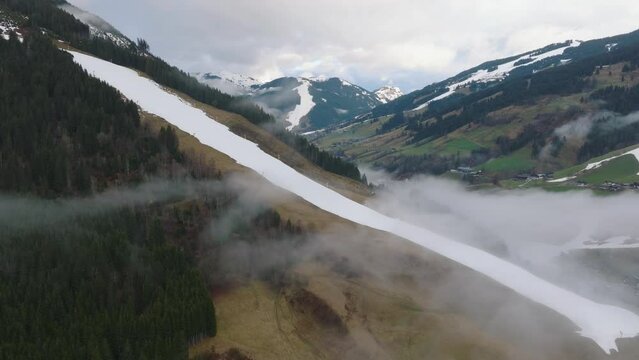 Mist over Saalbach-Hinterglemm (Austria) ski resort, snow-capped mountains flank a green valley, aerial view