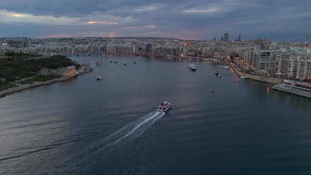 An aerial view of the Malta Sliema to Valletta ferry making its way into Sliema harbour at dusk, with the resort town in the background