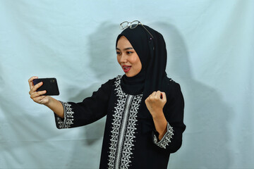 young Asian Muslim woman wearing hijab and black dress with white pattern using smartphone for...