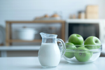 Pitcher of milk prepared for breakfast is placed near green apple bowl in the home kitchen, selective focus