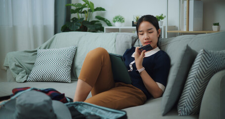 Portrait of Asian teenager woman sitting on sofa holding credit card making online payment on digital tablet, Preparation travel suitcase at home. - 772699347