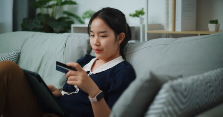 Selective focus of Asian teenager woman sitting on sofa holding credit card making online payment on digital tablet, Preparation travel suitcase at home. - 772699376