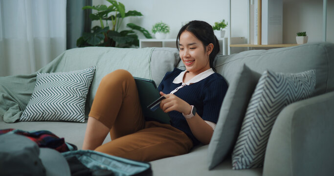 Portrait of Asian teenager woman sitting on sofa holding credit card making online payment on digital tablet, Preparation travel suitcase at home.