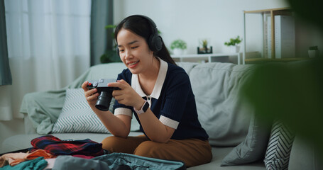 Selective focus of Asian teenager woman wear wireless headphones sitting on sofa look pictures on the camera while packing suitcase luggage in the living room , Preparation travel suitcase at home. - 772699512