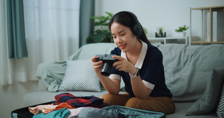 Selective focus of Asian teenager woman wear wireless headphones sitting on sofa look pictures on the camera while packing suitcase luggage in the living room , Preparation travel suitcase at home. - 772699519