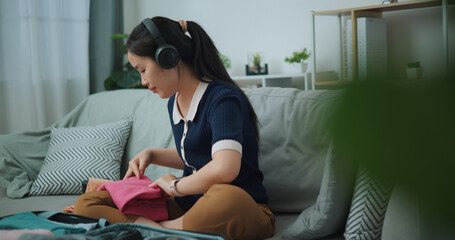Selective focus of Asian teenager woman wear wireless headphones sitting on sofa packing travel luggage with clothes for traveling trip, Preparation travel suitcase at home. - 772699534